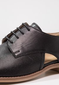 Chaussure noire en cuir avec un design perforé, texture lisse, couleur neutre et semelle en cuir beige. Comprend des lacets et des côtés ouverts.