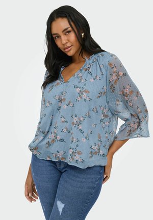 Kobieta z długimi ciemnymi włosami, ubrana w jasnoniebieską kwiecistą bluzkę i niebieskie dżinsy, pozująca z jedną ręką na biodrze, neutralne tło.