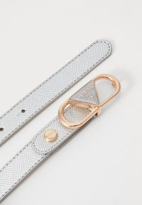 Curea texturată argintie cu un finisaj metalic, având o cataramă triunghiulară de culoare aurie, brănduită cu "GUESS". Stud metalic circular pe curea.