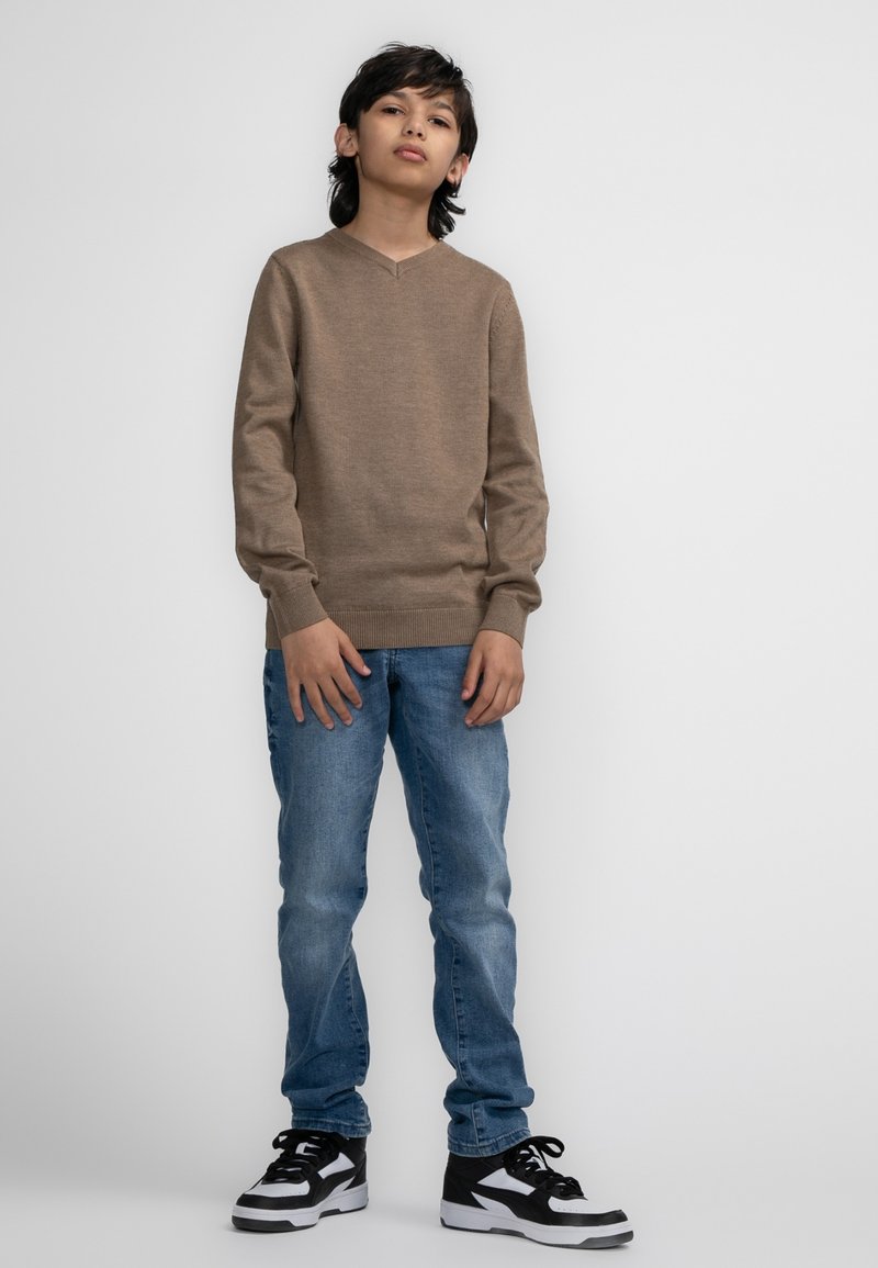 Beige V-Ausschnitt Pullover mit langen Ärmeln, kombiniert mit hellblauen Jeans und schwarzen Sneakers mit weißen Akzenten. Einfaches und lässiges Outfit.