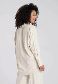 Lichtbeige oversized shirt met een kraag, opgerolde mouwen en een gebogen onderkant. Gelaagde stof met subtiele ontwerpdetails en gouden knoopaccenten.