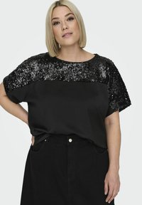 Czarna bluzka z krótkim rękawem, z ozdobnym górnym fragmentem pokrytym cekinami. Prosty design, gładka tekstura z błyszczącymi akcentami i luźny krój.
