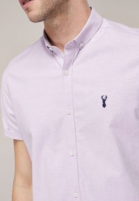 Camisa de manga corta de color púrpura claro con cuello, que presenta una tela texturizada y un pequeño logotipo bordado en azul oscuro. Los botones frontales son blancos.