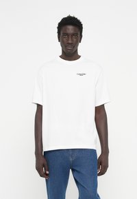 White cotton t-shirt with short sleeves, featuring a small black "Calvin Klein Jeans" logo on the chest, paired with blue denim jeans.