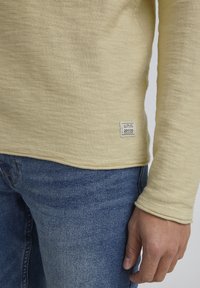 Personne portant un pull en maille jaune clair avec une petite étiquette et un jean en denim bleu, la main relâchée sur le côté, sur un fond neutre.