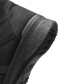 Semelle de chaussure en caoutchouc texturé noir avec dessus en daim et bandes en tissu cousues sur fond blanc, vue angulaire montrant le motif détaillé de la semelle.