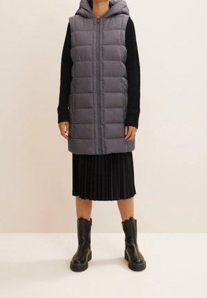 Mujer con chaleco tipo plumífero gris sin mangas con capucha, top de manga larga negro, falda negra plisada y botines negros, de pie contra un fondo liso.