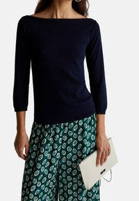 Marineblaues Bootsausschnitt-Pullover mit Dreiviertelärmeln, kombiniert mit grünen gemusterten Weitbeinhosen und einer weißen strukturierten Clutch.