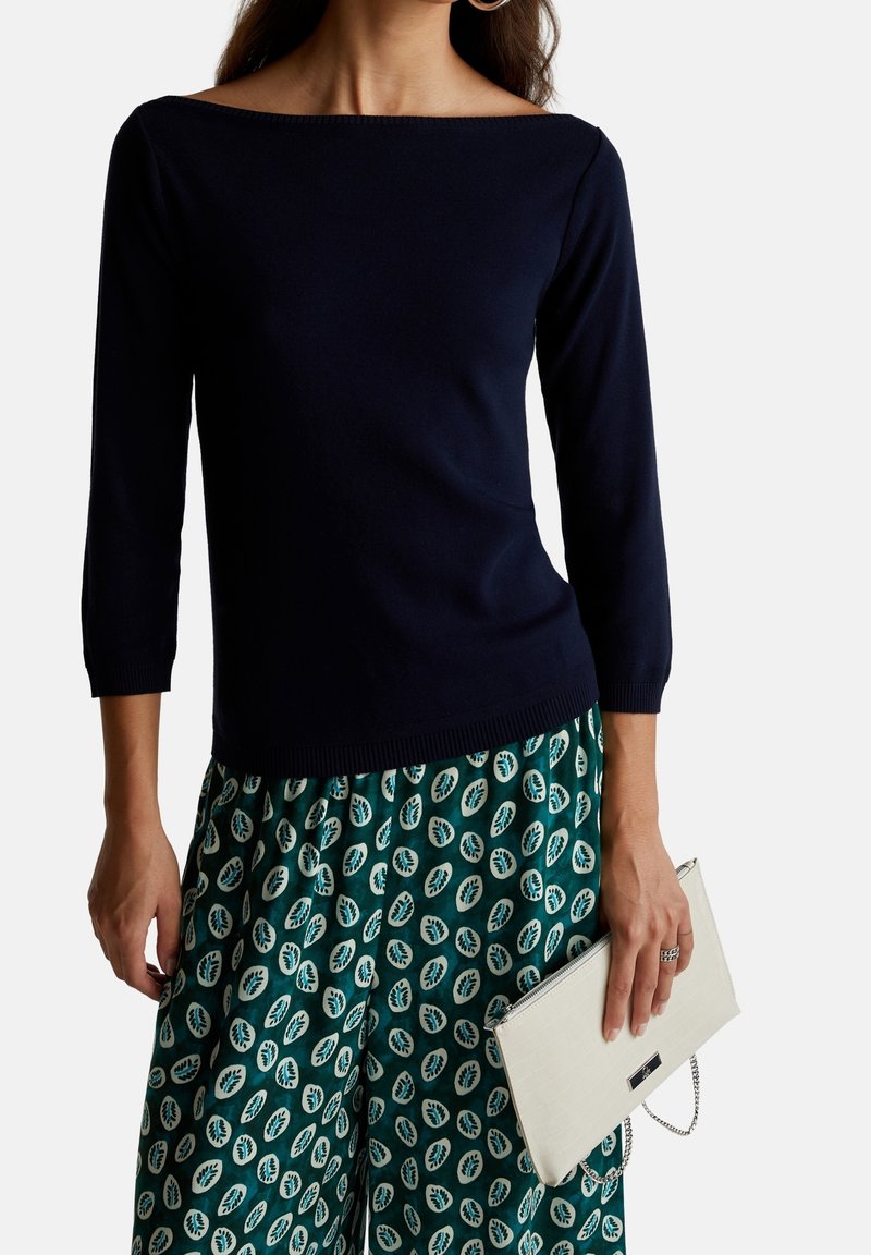 Marineblaues Bootsausschnitt-Pullover mit Dreiviertelärmeln, kombiniert mit grünen gemusterten Weitbeinhosen und einer weißen strukturierten Clutch.