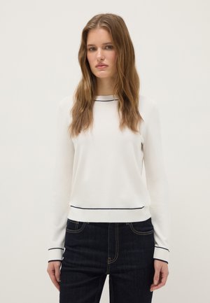 Joven con cabello largo castaño claro que lleva una blusa blanca de manga larga con ribetes negros y jeans azul oscuro, de pie frente a un fondo blanco liso.