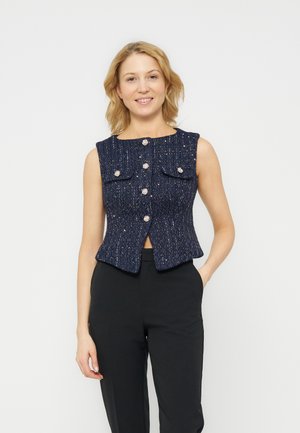 Nainen hymyilee pukeutuneena hihattomaan laivastonsiniseen tweeddiliiviin, jossa on koristeelliset napit, ja mustiin housuihin, seisten käsi taskussa.