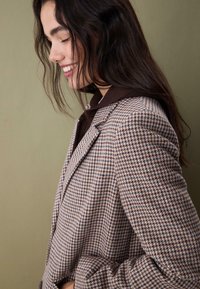 Brauner Houndstooth-Blazer mit strukturiertem Design, Reverskragen und strukturiertem Stoff, über einem dunkelbraunen Oberteil gestapelt.