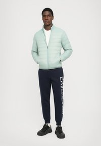 Jachetă puffer verde deschis cu fermoar frontal și buzunare laterale, asortată cu pantaloni de jogging bleumarin închis, având text grafic alb. Sneakers negri.