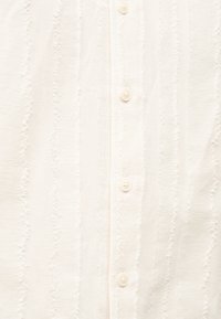 Ύφασμα σε εκρού με υφή και κάθετες κυματιστές ρίγες και μικρά στρογγυλά κουμπιά στη μέση.