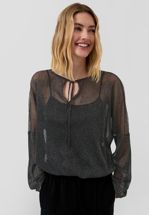 Jonge vrouw die glimlacht en een transparante zwarte glinsterende blouse met een strik aan de voorkant over een zwarte camisole draagt, met haar handen in de zakken van een zwarte broek.