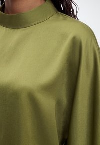 Gros plan d'une personne portant un haut lisse vert olive à col montant avec un tissu doux drapé sur l'épaule.