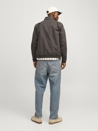 Black bomber jacket with a stand collar, light blue jeans, and beige slip-on shoes. The model wears a white cap with a black brim.