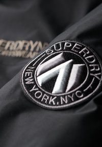 Sort stof med et fremtrædende broderet cirkulært logo med hvide og grå accenter, der står "Superdry New York, NYC." Glat tekstur.