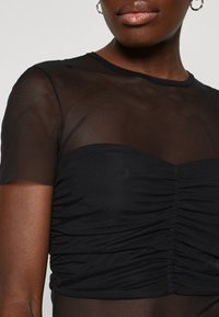 Top negro transparente con mangas cortas, que presenta una zona de busto fruncida con textura suave, destacando un diseño en capas y una forma ajustada.