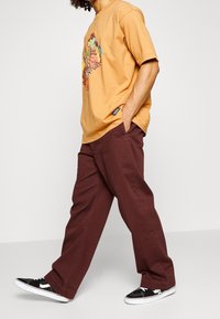 Bruna bomullbyxor med lös passform, tillsammans med en orange grafisk t-shirt med färggrafik. Svarta sneakers med vita sulor.