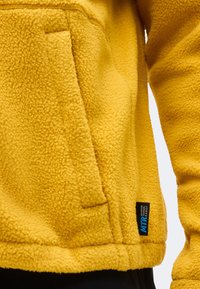 Veste en polaire jaune avec une poche avant et une étiquette noire indiquant « MTR Mountain Technical Range » sur la partie inférieure.