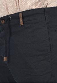 Pantalon en tissu noir avec une taille à cordon de serrage, doté d'une étiquette de marque en cuir marron et d'une texture lisse, présenté de profil.