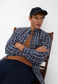 Gekruiste blauw-witte overhemd over een bruine t-shirt, gecombineerd met marineblauwe broek; dragend een zwarte pet met blauwe rand, zittend op een houten stoel.