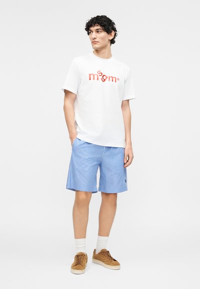 Νεαρός άνδρας φορώντας λευκό μπλουζάκι με στάμπα, μπλε και λευκή ριγέ βερμούδα, λευκές κάλτσες και καφέ σουέτ αθλητικά παπούτσια, στέκεται με τα χέρια στις τσέπες.