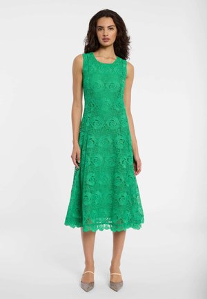 Frau trägt ein ärmelloses, grünes Spitzen-Midikleid mit Blumenmustern und beige spitze Absätze, stehend vor einem schlichten weißen Hintergrund.