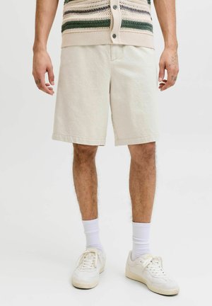 Mężczyzna w beżowych szortach do kolan, białych trampkach, białych skarpetkach i bezrękawniku wykonanym z dzianiny w zielone i beżowe paski.