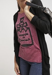 Bordová tričko s grafikou parfémové lahvičky zdobené černými flitry, v kombinaci s černým kardiganem s otevřeným úpletem. Na sobě má modré džíny a stříbrnou tašku.