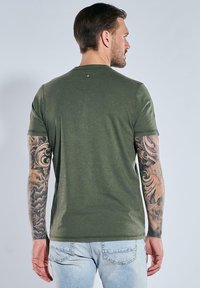 Olive green cotton t-shirt with short sleeves, crew neckline, and a subtle back seam detail. Notable tattoos visible on arms.