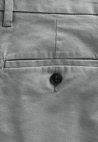 Tissu gris avec une texture fine, doté d'une fermeture à boutons et d'une petite poche. Le design est épuré et minimaliste.