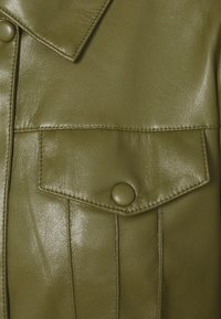 Gros plan d'une veste en cuir vert olive, présentant une poche à rabat boutonnée, des coutures visibles et une texture lisse avec une finition mate.