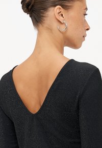Čierny dlhý rukáv top s výstrihom do V na chrbte; trblietavá textúra zvyšuje eleganciu. Model má na sebe strieborné kruhové náušnice.