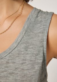 Siv brezrokavnik iz mehke, teksturirane tkanine. Ima globok izrez in neravne robove, dopolnjen z večplastno zlato verižico.