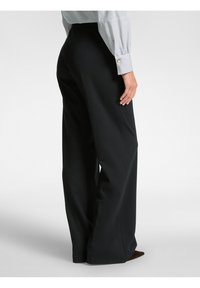 Vrouw in hoge, wijde zwarte broek met een witte blouse met lange mouwen en zwarte strepen, staande van opzij.