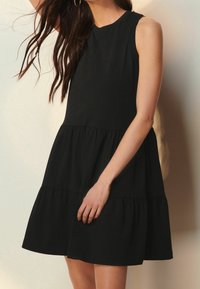 Czarna sukienka bez rękawów z okrągłym dekoltem, zaprojektowana w warstwowym stylu, z miękką fakturą materiału i luźnym krojem, sięgająca powyżej kolan.