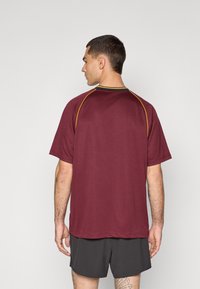 Bordeauxkleurig sportshirt met korte mouwen, voorzien van groene en gele accenten rond de kraag en mouwen, met een gestructureerde stof.