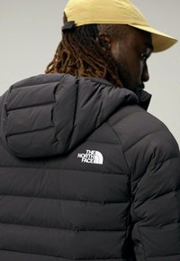 Giacca a vento nera isolata con cappuccio e logo sulla spalla sinistra. Il tessuto sembra liscio e leggermente lucido.