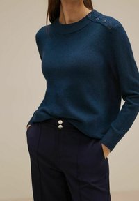 Marinblå stickad tröja med rundad halsringning, långa ärmar och fyraknapparsdetalj på axeln. Matchas med höga marinblå byxor.