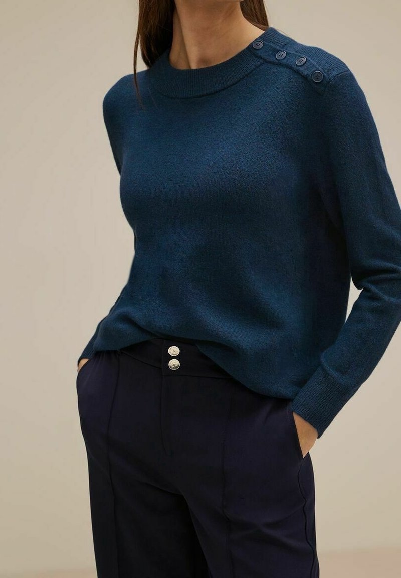 Marinblå stickad tröja med rundad halsringning, långa ärmar och fyraknapparsdetalj på axeln. Matchas med höga marinblå byxor.