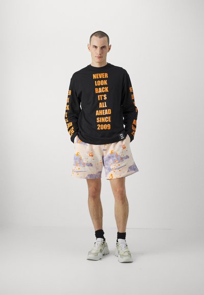 Chemise noire à manches longues avec un texte orange en gras, assortie à un short crème présentant des motifs éclaboussés de violet et d'orange. Baskets grises et chaussettes noires.