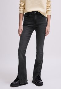 Pantalones vaqueros negros acampanados con aberturas delanteras, hechos de mezclilla. Cuenta con una cintura alta y diseño de cinco bolsillos, combinados con botas negras de suela gruesa.