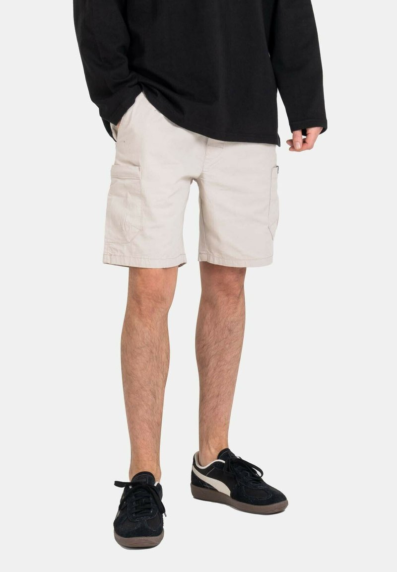 Pantalones cortos cargo beige con dos bolsillos laterales, hechos de algodón. Combinados con zapatillas negras con suela y cordones de color marrón claro.