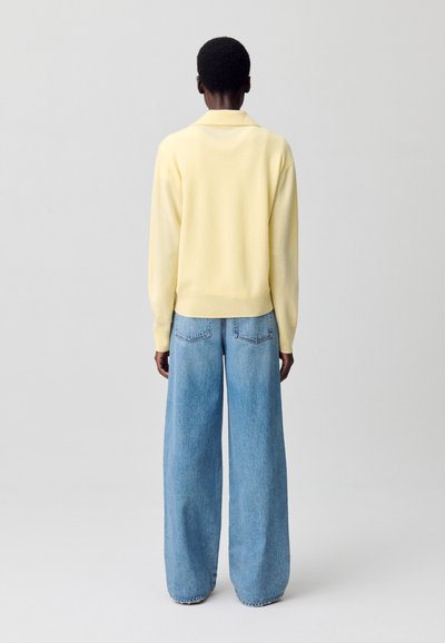 Suéter amarillo de punto ligero con cuello, combinado con pantalones vaqueros de denim azul claro de pierna amplia; costuras visibles y bolsillos traseros.