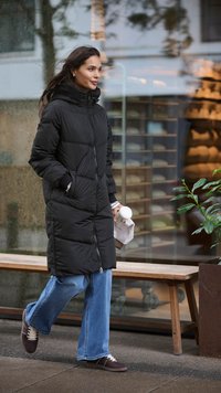 Cappotto imbottito nero con cappuccio, maniche lunghe e texture trapuntata, abbinato a jeans larghi blu e sneakers scure, mentre tiene in mano una tazza di caffè.
