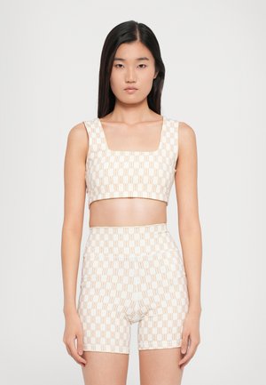 Young woman with straight black hair wearing a beige crop top and matching high-waisted shorts with a repeating logo pattern.