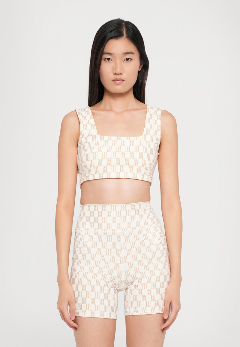 Young woman with straight black hair wearing a beige crop top and matching high-waisted shorts with a repeating logo pattern.