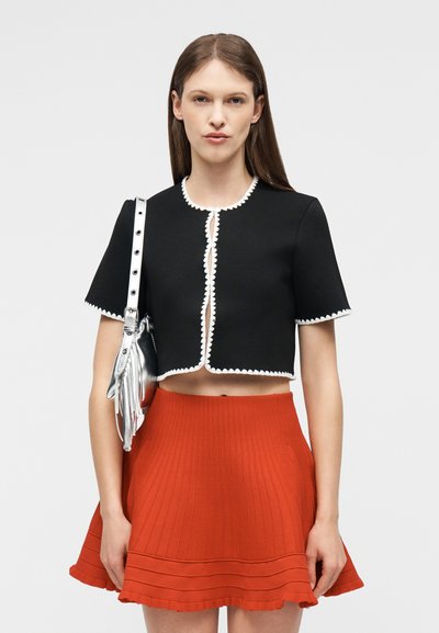 Νεαρή γυναίκα που φοράει μαύρο cropped μπουφάν με λευκή επένδυση, κόκκινη πλισέ φούστα και κρατάει μια μεταλλική ασημένια τσάντα στον ώμο της.